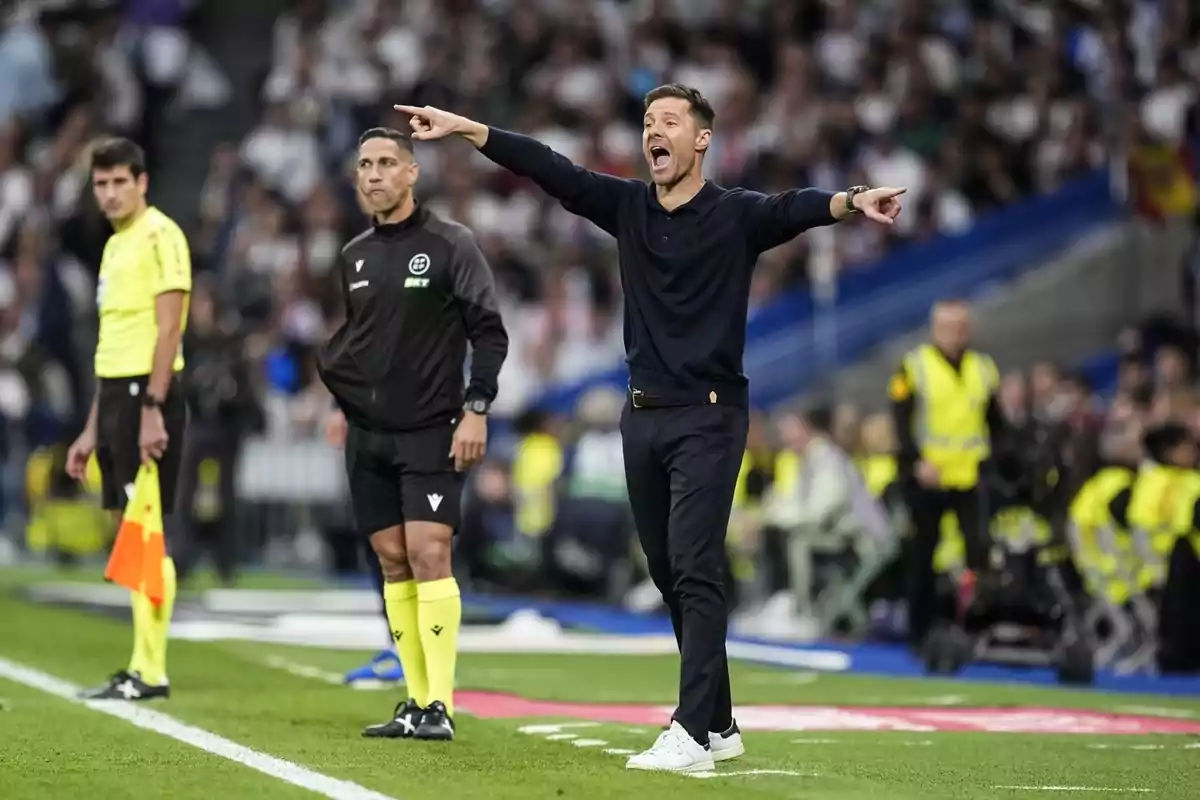 Entrenador de futbol donant indicacions a la línia de banda durant un partit amb àrbitres i assistents al camp i públic a les grades.