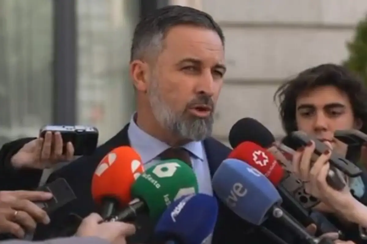 Un hombre de barba y cabello corto habla frente a varios micrófonos de diferentes medios de comunicación mientras es rodeado por periodistas