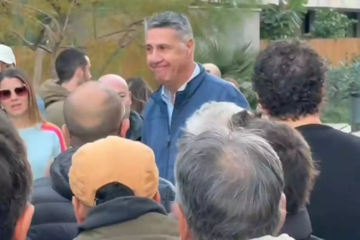 Un hombre de cabello canoso y chaqueta azul está rodeado de varias personas al aire libre en un ambiente urbano con vegetación al fondo