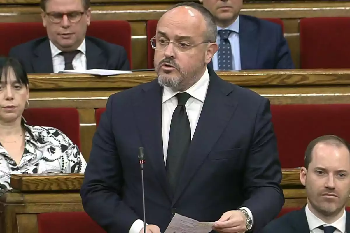 Hombre de traje oscuro y corbata negra hablando en un parlamento con varias personas sentadas detrás de él