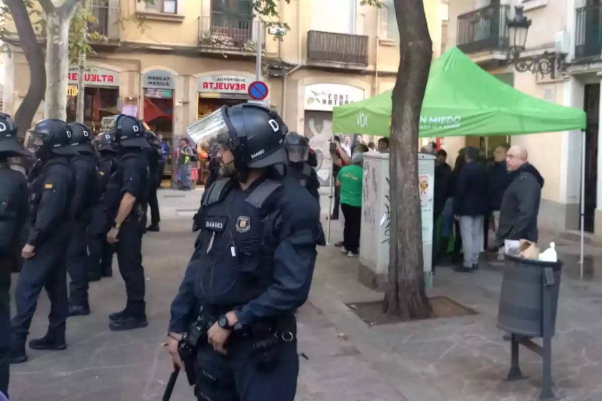 Diversos policies antidisturbis vigilen una plaça on hi ha una carpa verda i diverses persones reunides a prop d’un edifici antic.