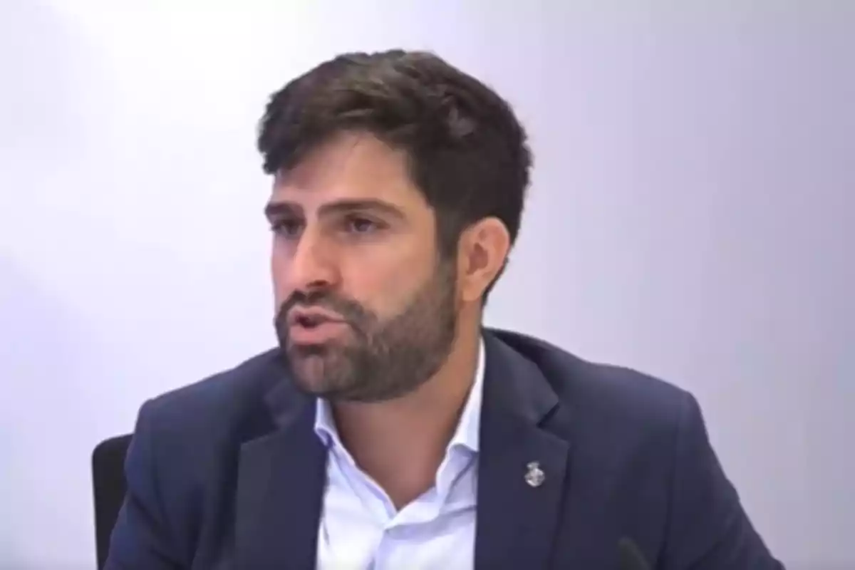 Home de cabells foscos i barba parlant mentre porta una americana blau marí i una camisa blanca en un entorn professional