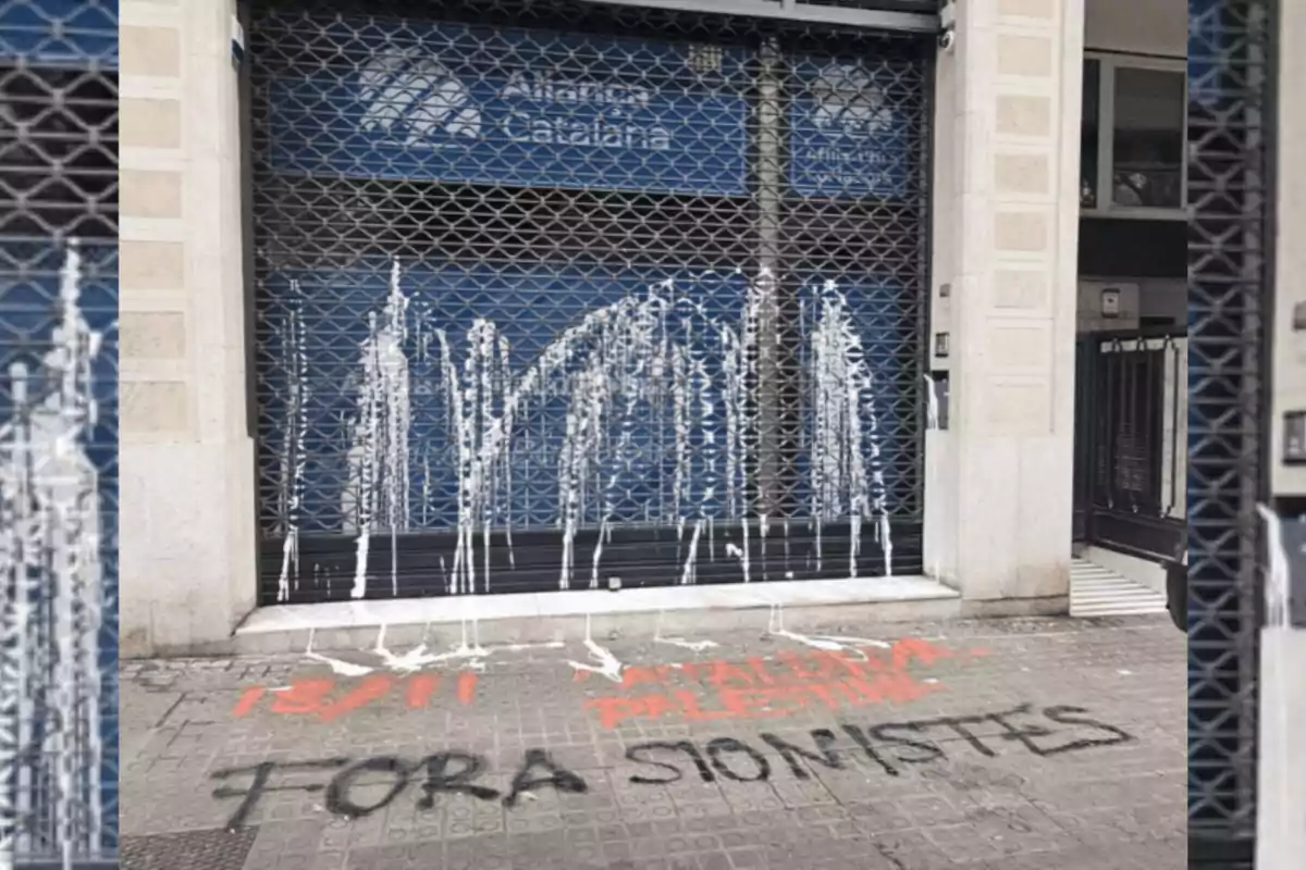 Fachada de un local con una reja metálica manchada de pintura blanca y pintadas en el suelo con mensajes en catalán y español, incluyendo una frase en negro que dice "FORA SIONISTES"