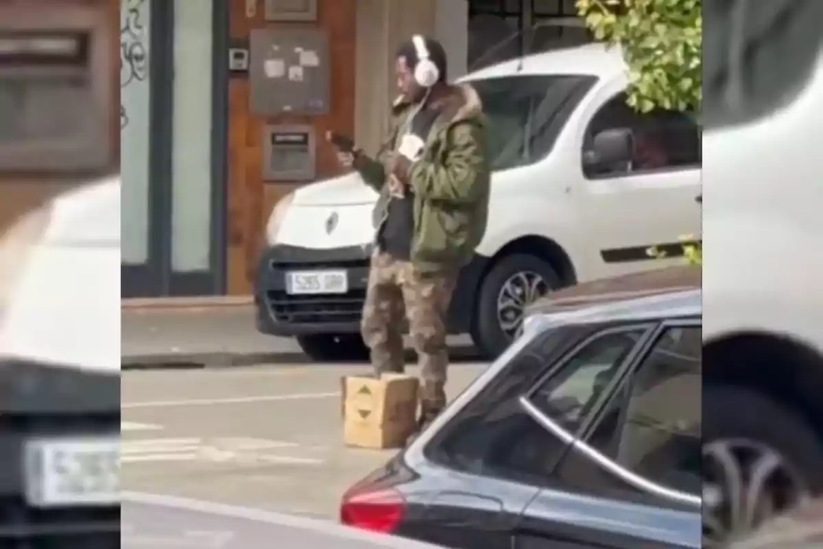 Hombre con auriculares de pie en la calle sosteniendo un teléfono móvil y una bebida, vestido con chaqueta verde y pantalones de camuflaje, junto a una caja de cartón y varios autos estacionados