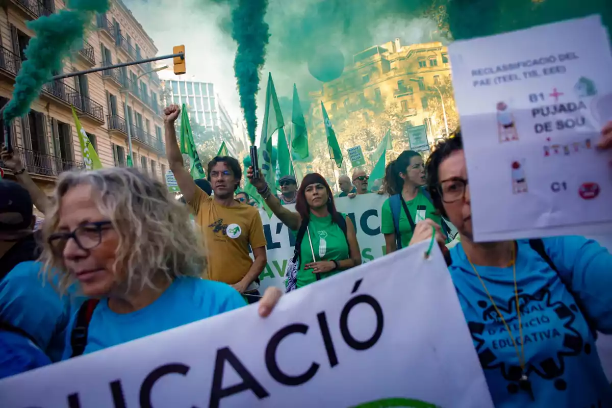 Personas marchando en una manifestación educativa con pancartas, camisetas azules y verdes, y humo verde en el aire en una calle de ciudad