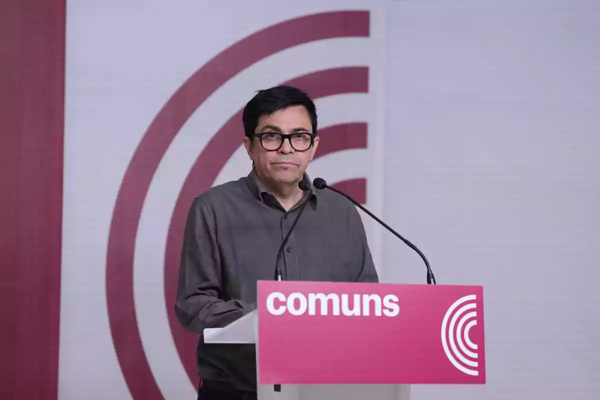 Hombre con gafas y camisa gris hablando en un atril con el logo y la palabra comuns sobre fondo blanco y burdeos