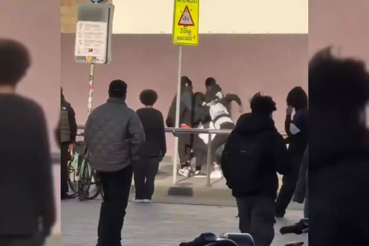 Varias personas observan una pelea en la vía pública cerca de una señal de zona escolar.