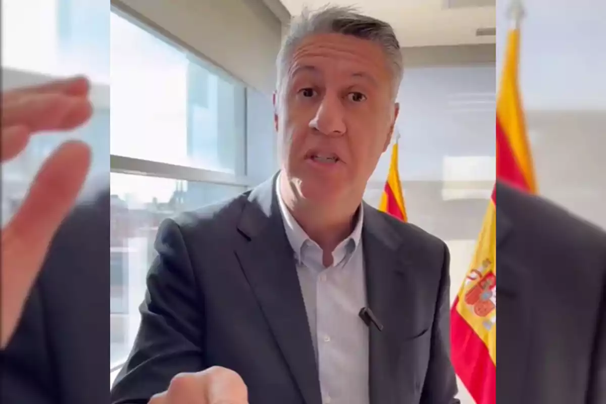 Hombre de cabello canoso con traje oscuro hablando frente a una ventana y dos banderas de España al fondo