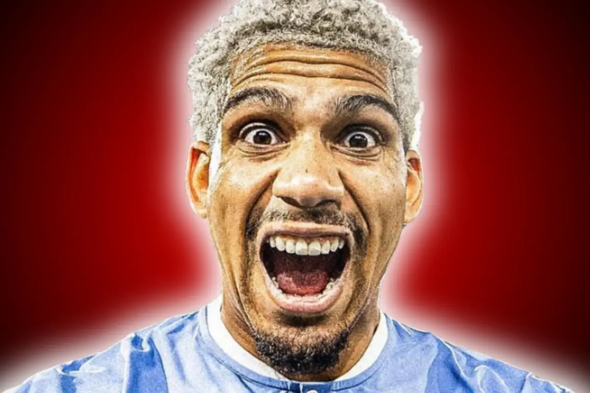 Hombre de cabello rizado y canoso con expresión de sorpresa o emoción, vistiendo una camiseta azul y fondo rojo difuminado.