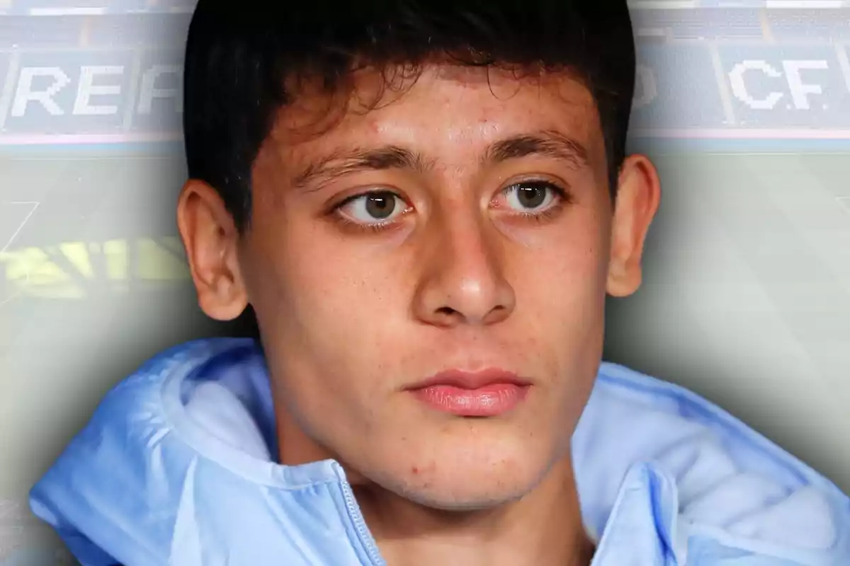 Young man in a light blue jacket looking straight ahead with a serious expression and a blurred football stadium background