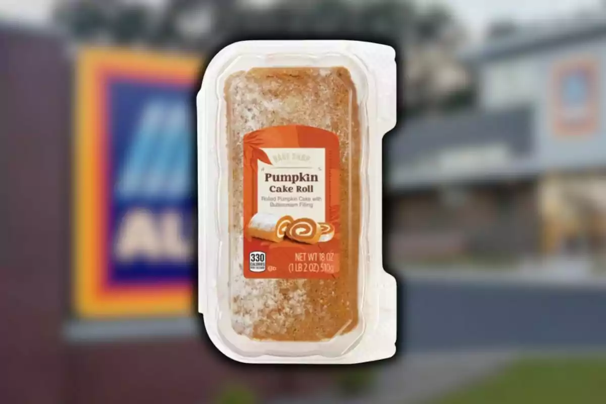 Un rollo de pastel de calabaza en empaque transparente con una etiqueta que indica que es de la marca Bake Shop y fondo borroso de una tienda Aldi.