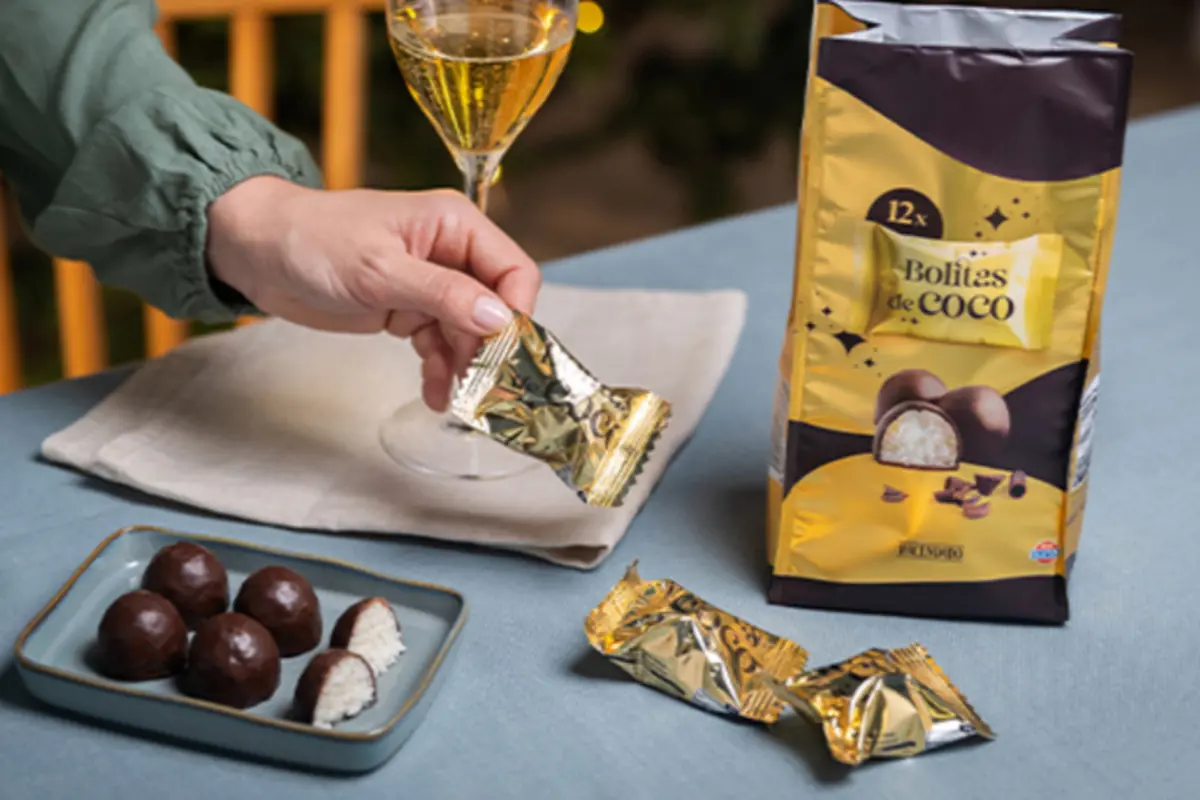 Una mano sostiene un dulce envuelto en papel dorado junto a una copa de vino, un plato con bolitas de coco cubiertas de chocolate y una bolsa amarilla de bolitas de coco sobre una mesa azul
