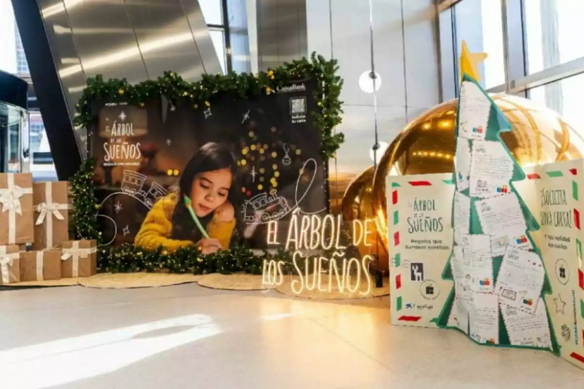 Decoració nadalenca en un espai interior amb regals embolicats, un cartell d’una nena escrivint, un rètol lluminós que diu L’Arbre dels Somnis i un arbre fet de cartes al costat d’una esfera daurada gegant.