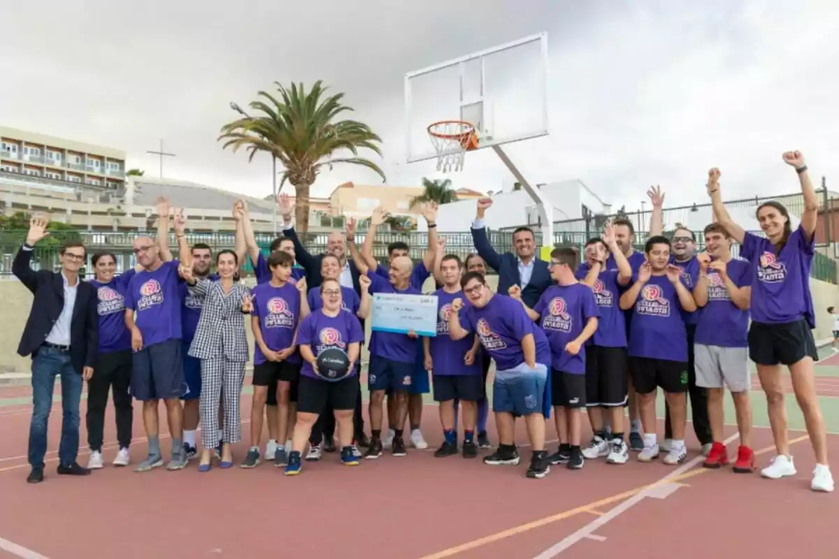 Grup de persones amb samarretes liles celebrant en una pista de bàsquet a l’aire lliure, alguns aixequen els braços i un sosté una pilota i un altre un xec gran.