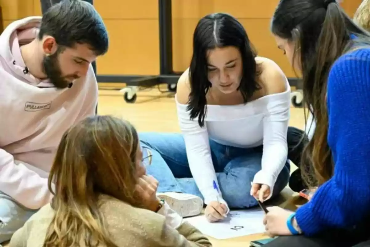 Quatre joves asseguts a terra treballen junts escrivint en un full de paper durant una activitat grupal a una aula. Quatre joves asseguts a terra treballen junts escrivint en un full de paper durant una activitat grupal a una aula.
