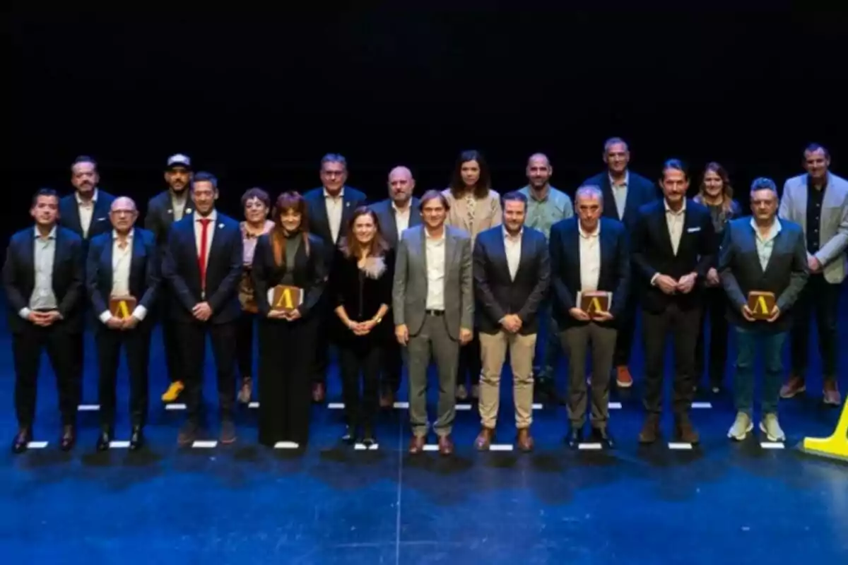 Grup de persones posant en un escenari, algunes d'elles sostenen plaques o premis, tots vestits de manera formal i amb un fons fosc.
