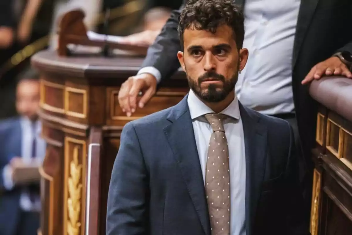 Hombre de cabello rizado y barba con traje y corbata marrón de lunares en un entorno parlamentario Hombre de cabello rizado y barba con traje y corbata marrón de lunares en un entorno parlamentario