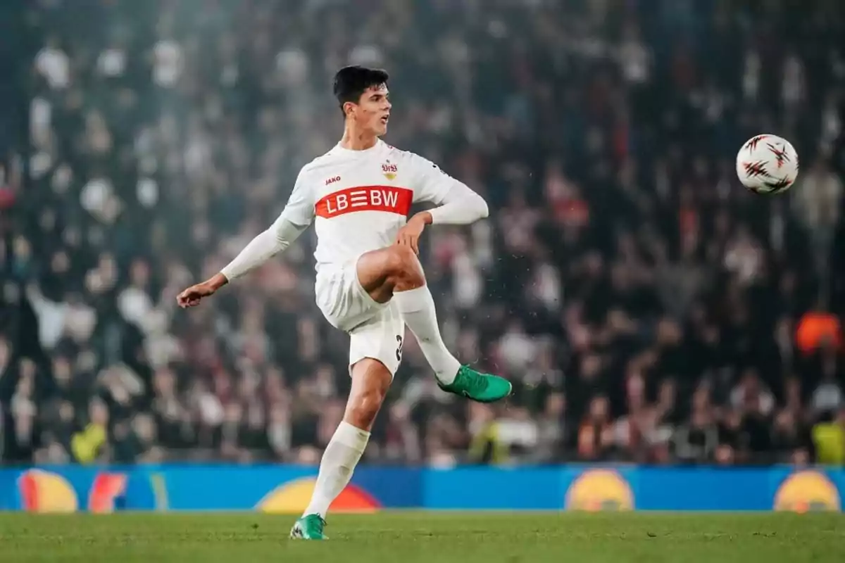 Futbolista amb uniforme blanc i franja vermella xutant una pilota en un estadi ple d’espectadors