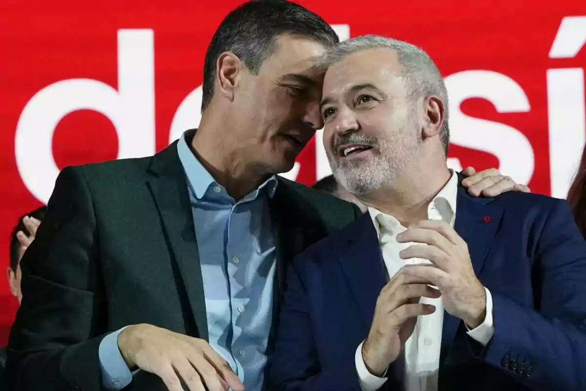 Dos hombres vestidos de traje conversan sonrientes frente a un fondo rojo con letras blancas Dos hombres vestidos de traje conversan sonrientes frente a un fondo rojo con letras blancas