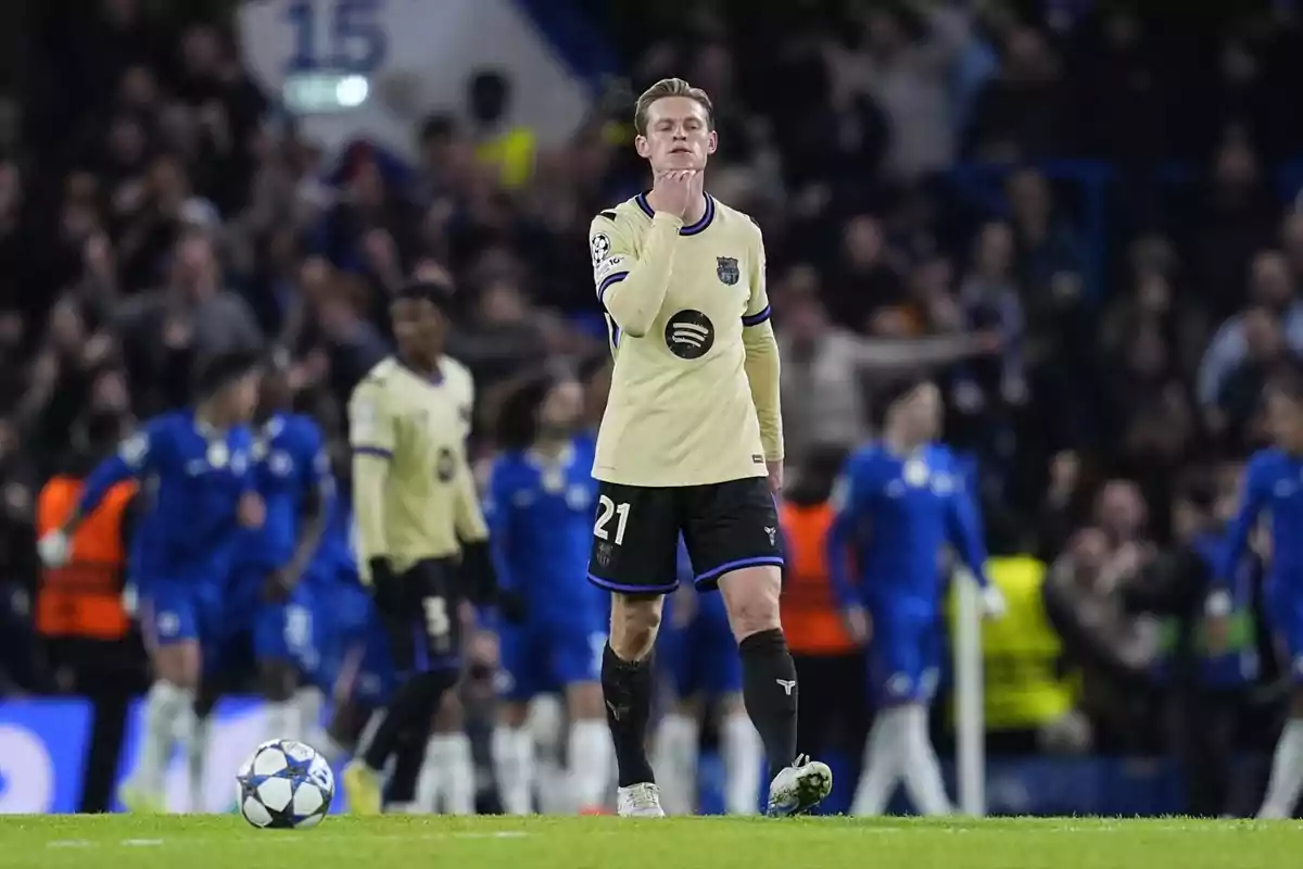 Futbolista con uniforme crema y negro caminando pensativo en el campo durante un partido mientras otros jugadores y público se ven desenfocados al fondo Futbolista con uniforme crema y negro caminando pensativo en el campo durante un partido mientras otros jugadores y público se ven desenfocados al fondo