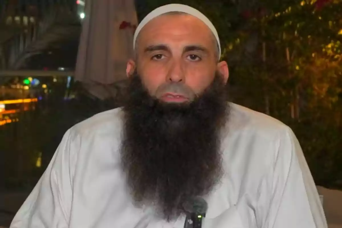 Home amb barba llarga i gorra blanca assegut a l’aire lliure amb vegetació de fons