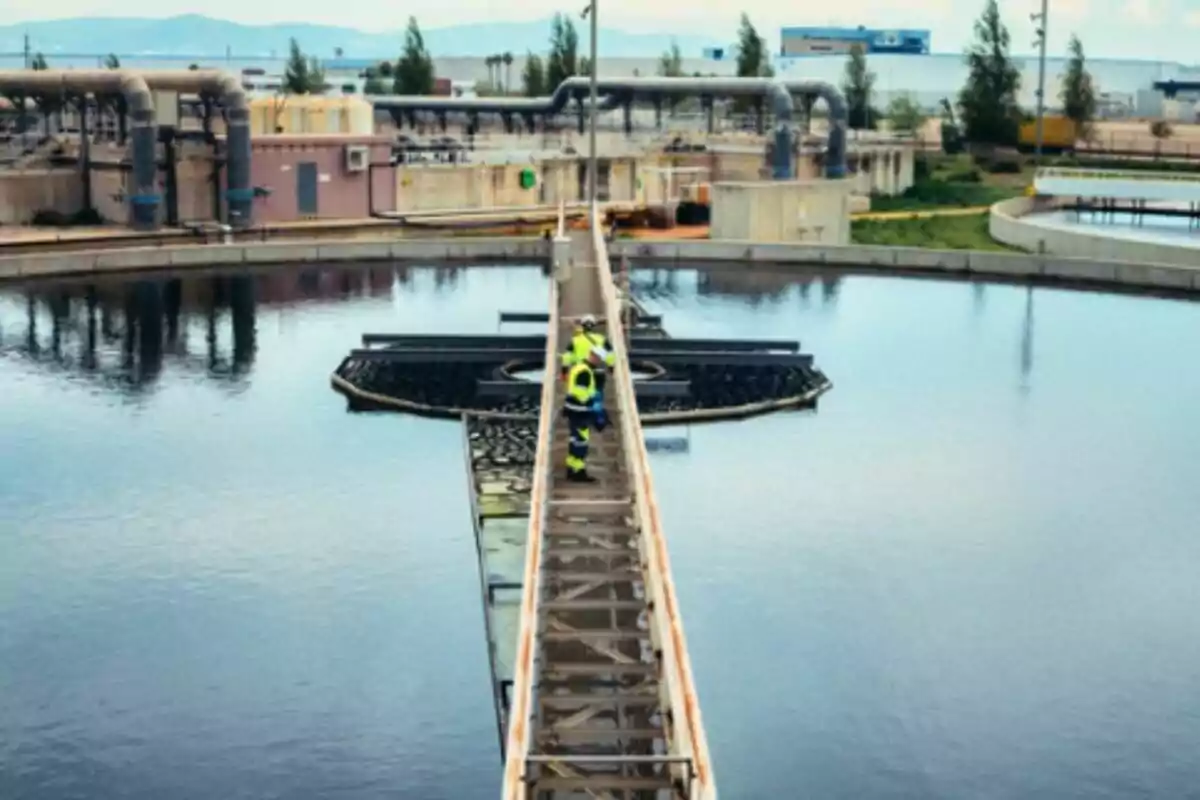 Trabajador con chaleco de seguridad caminando sobre una pasarela en una planta de tratamiento de aguas rodeada de tanques y tuberías industriales.
