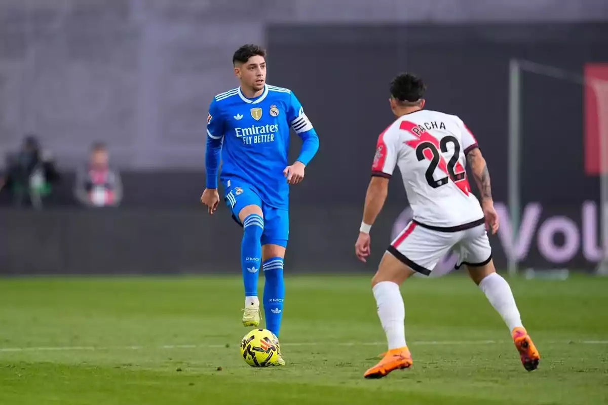 Un jugador del Reial Madrid amb uniforme blau controla la pilota mentre un jugador del Rayo Vallecano amb uniforme blanc i detalls vermells el marca durant un partit de futbol al camp.