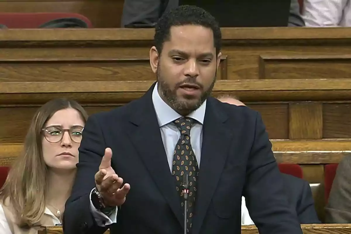 Hombre de traje hablando en un parlamento con una mujer sentada detrás observando atentamente