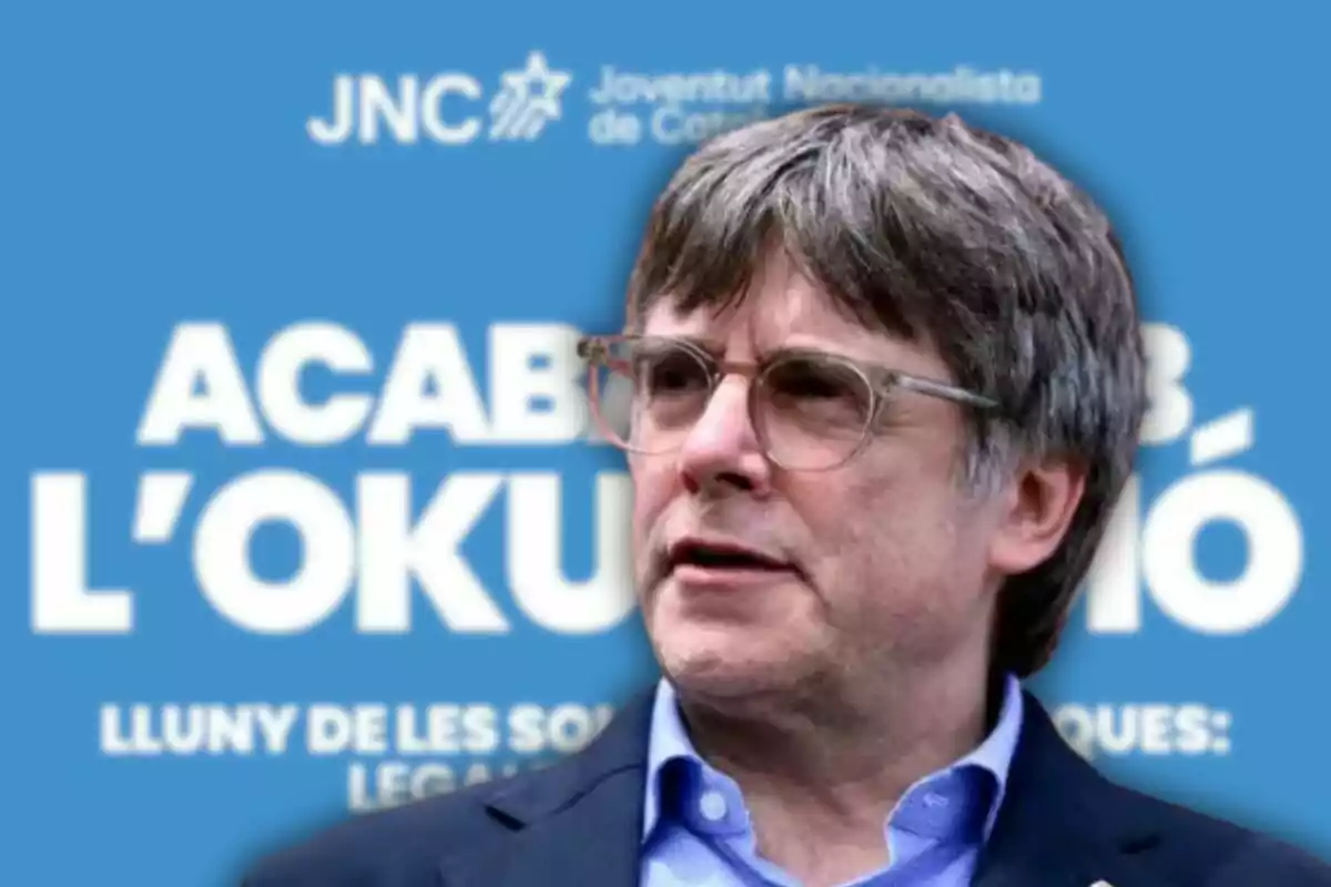 Hombre de cabello canoso y gafas frente a un cartel azul con texto en catalán y el logo de la Joventut Nacionalista de Catalunya.