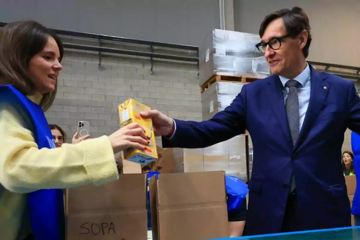 Dos personas colaboran en el embalaje de alimentos en una bodega, una de ellas entrega un cartón de sopa a la otra frente a cajas de cartón y palets apilados.