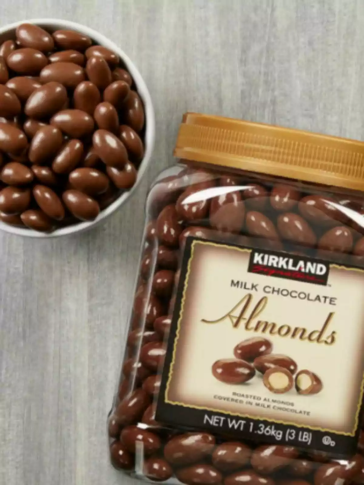 Jar of milk chocolate-covered almonds from Costco next to a white bowl filled with the same product on a gray table. Jar of milk chocolate-covered almonds from Costco next to a white bowl filled with the same product on a gray table.