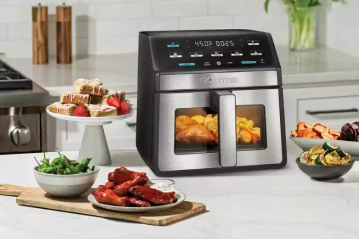 Costco stainless steel air fryer in a modern kitchen surrounded by various dishes such as wings, vegetables, peppers, and desserts with strawberries.