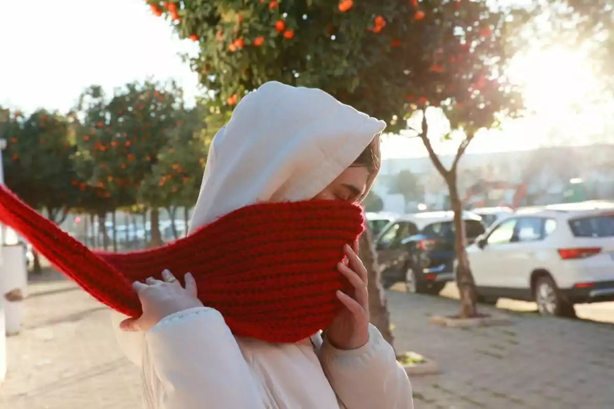 Persona amb abric blanc i bufanda vermella cobrint-se el rostre mentre camina per un carrer amb arbres i cotxes aparcats al capvespre.