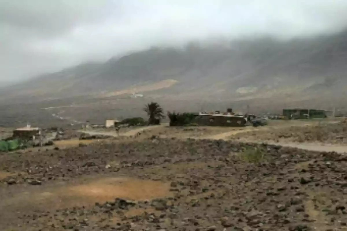Paisatge àrid amb sòl rocós, algunes cases disperses i muntanyes cobertes de boira al fons a Canàries.