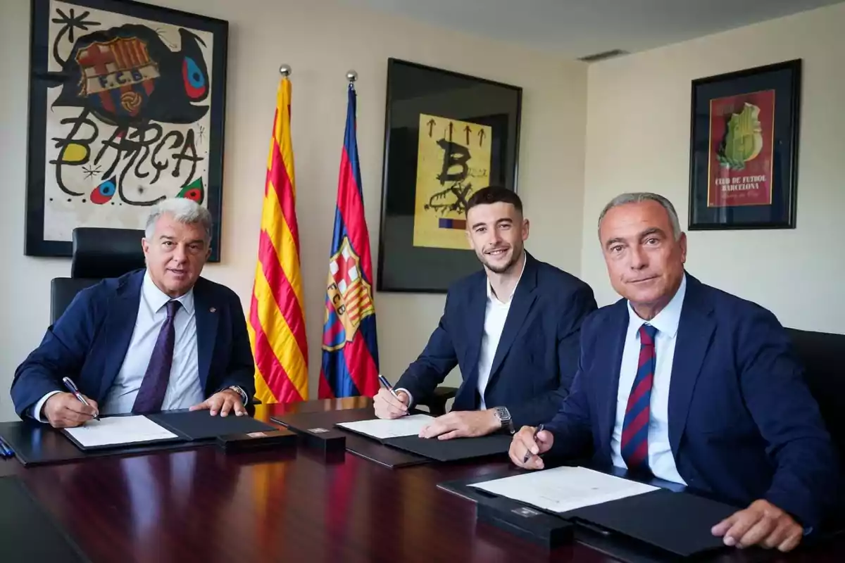 Tres homes amb vestits formals asseguts en una taula signant documents amb banderes i quadres del FC Barcelona al fons