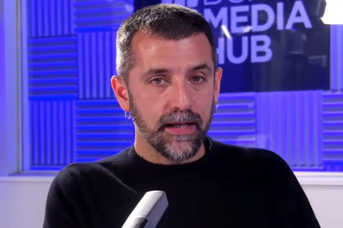 Hombre de barba y cabello corto hablando frente a un micrófono en un estudio con paredes de paneles acústicos azules y un letrero de fondo