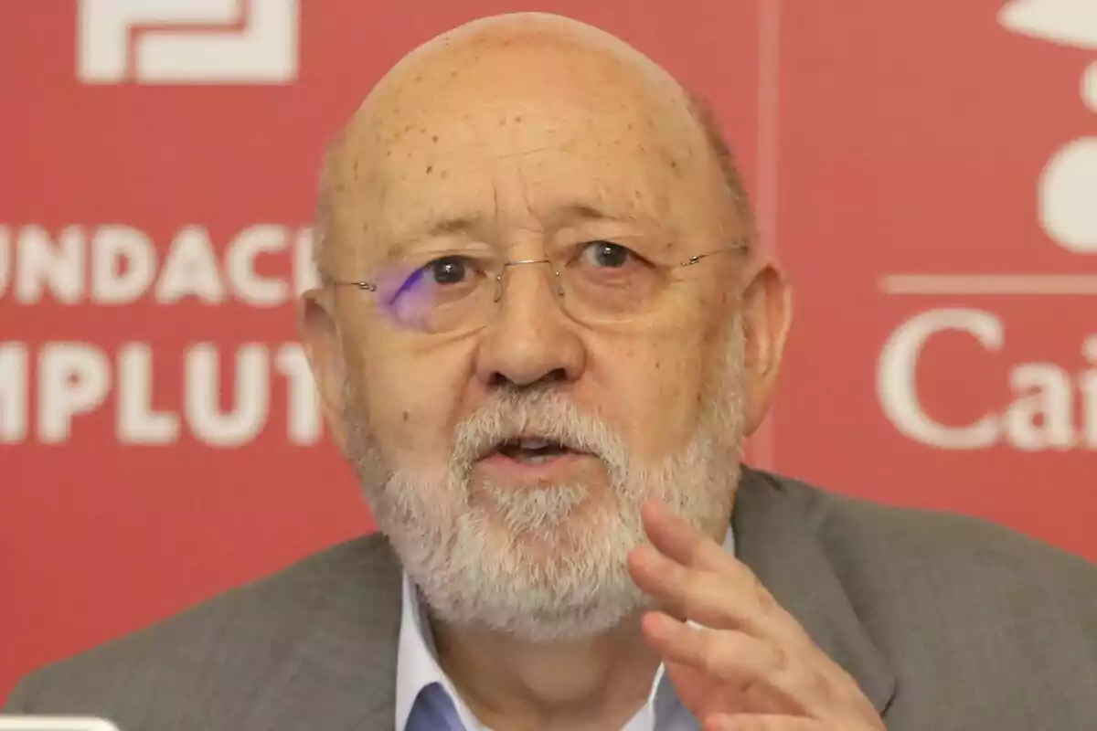 José Félix Tezanos amb barba i ulleres parlant davant d’un fons vermell amb lletres blanques.