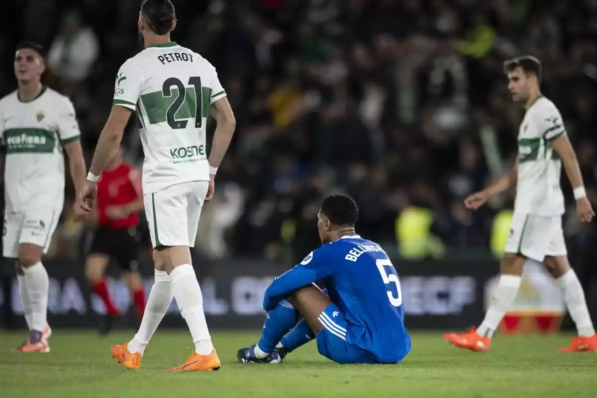 Un jugador con uniforme azul está sentado en el césped durante un partido de fútbol mientras varios jugadores con uniforme blanco caminan a su alrededor