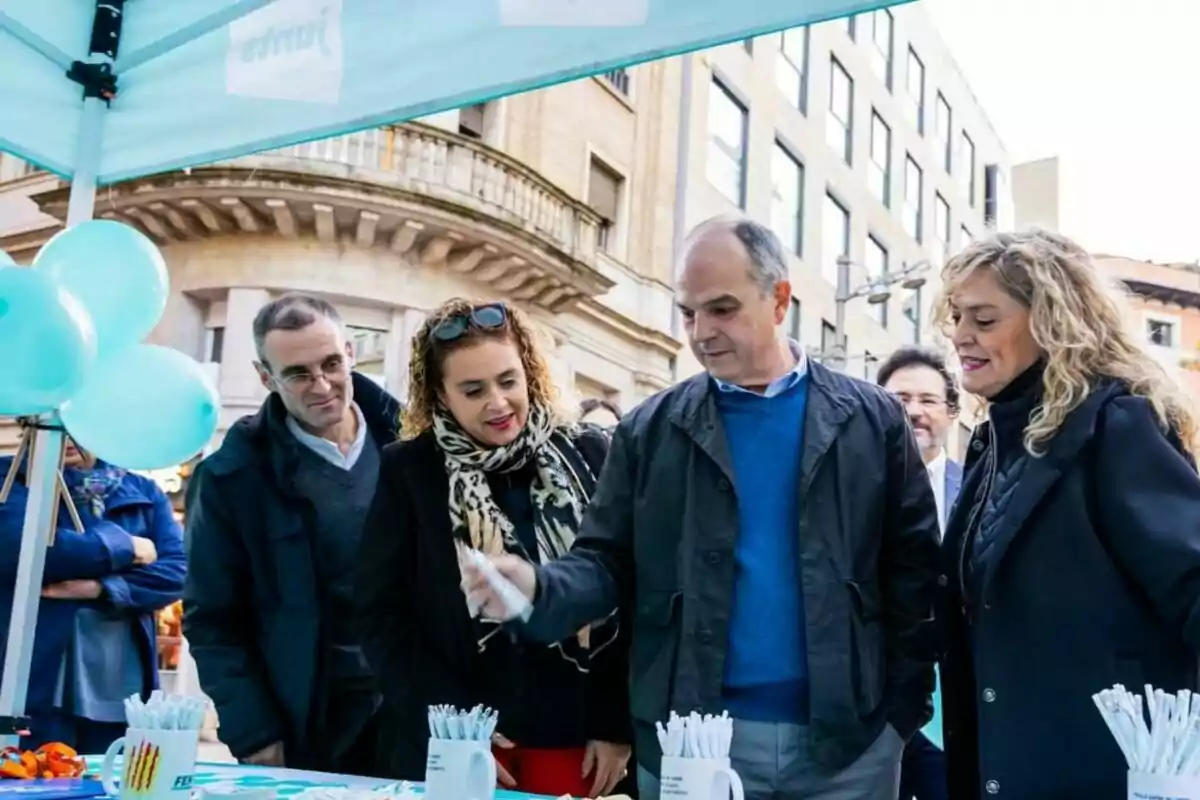 Varias personas observan una mesa con bolígrafos y tazas promocionales bajo una carpa en un evento al aire libre en la ciudad