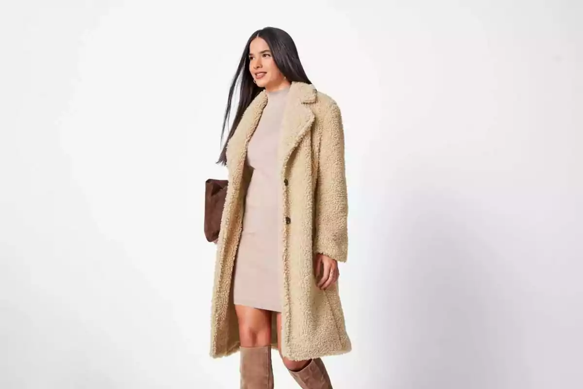 Woman in a beige shearling coat, light dress, and tall brown boots, holding a dark handbag against a white background