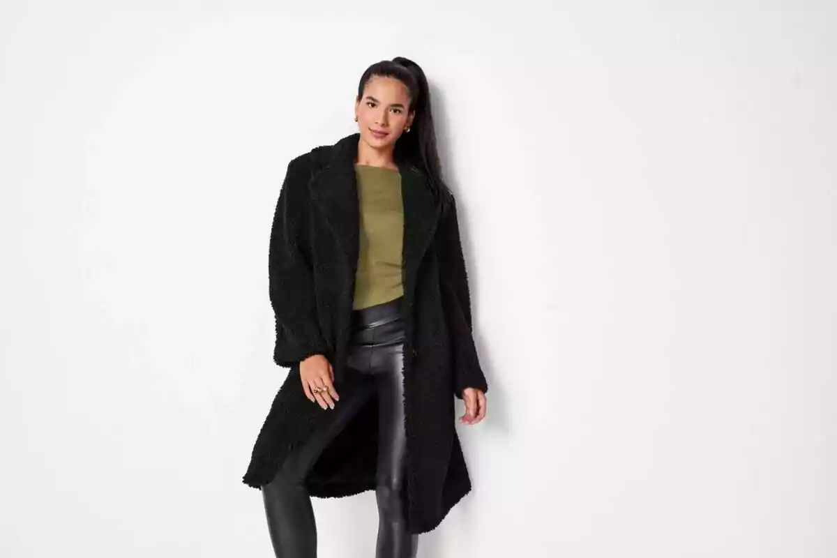 Young woman in a long black coat, green blouse, and black leather pants posing in front of a white wall
