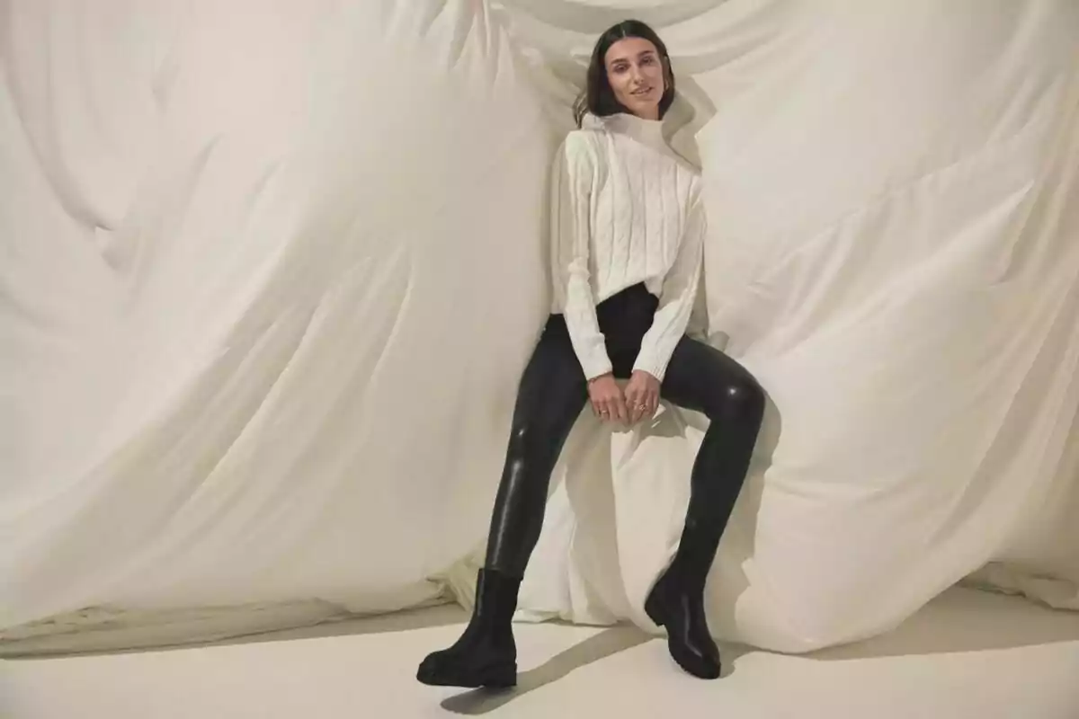 Woman posing in a white knit sweater, black leggings, and black boots in front of a beige fabric backdrop Woman posing in a white knit sweater, black leggings, and black boots in front of a beige fabric backdrop