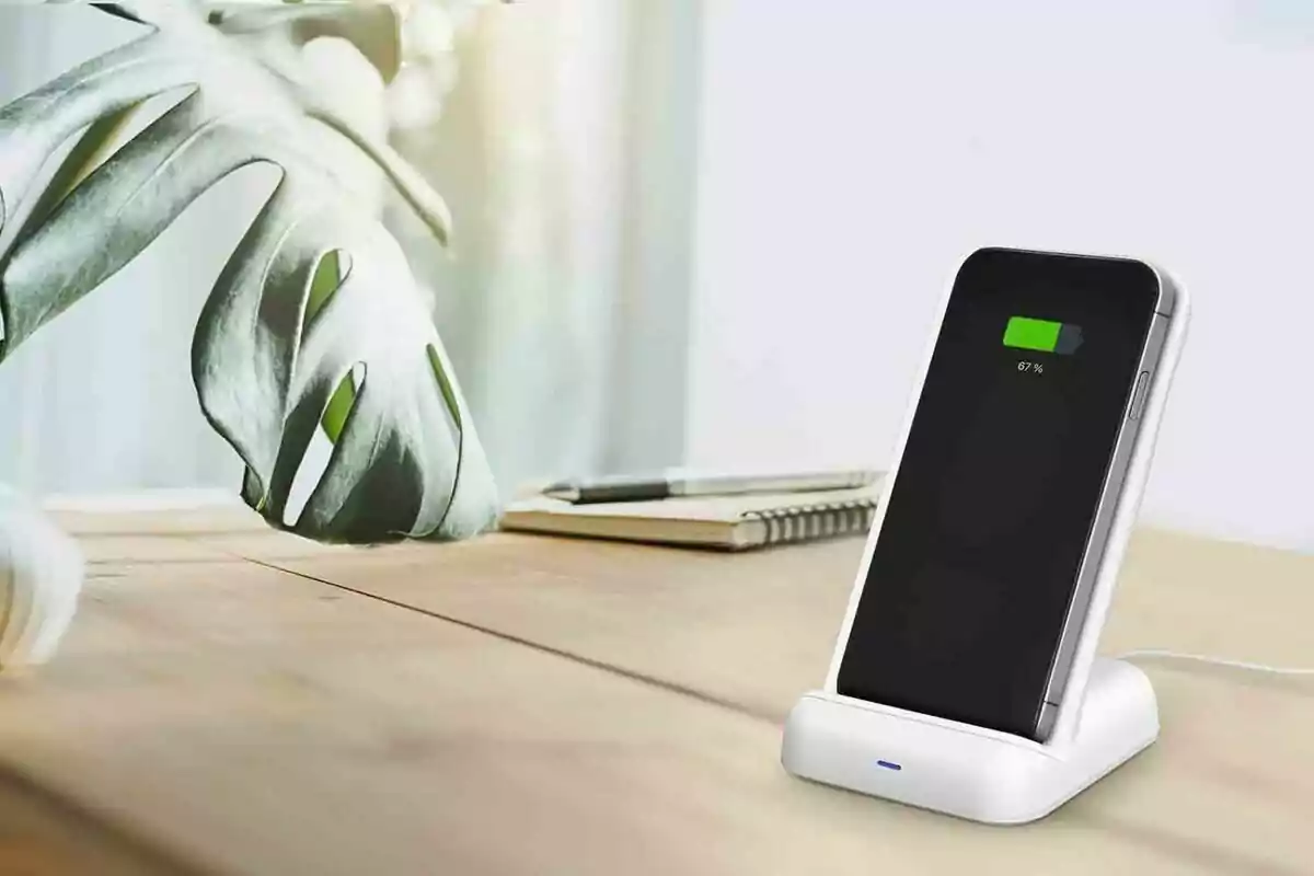 Mobile phone charging on a wireless base on a wooden desk next to a notebook and a decorative plant