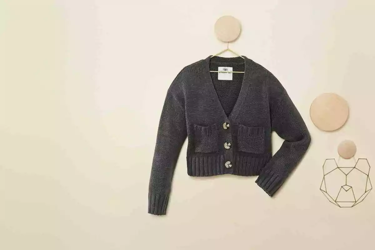 Dark gray knit sweater with large buttons and two front pockets hanging on a beige wall decorated with wooden geometric shapes and a metal bear-shaped ornament