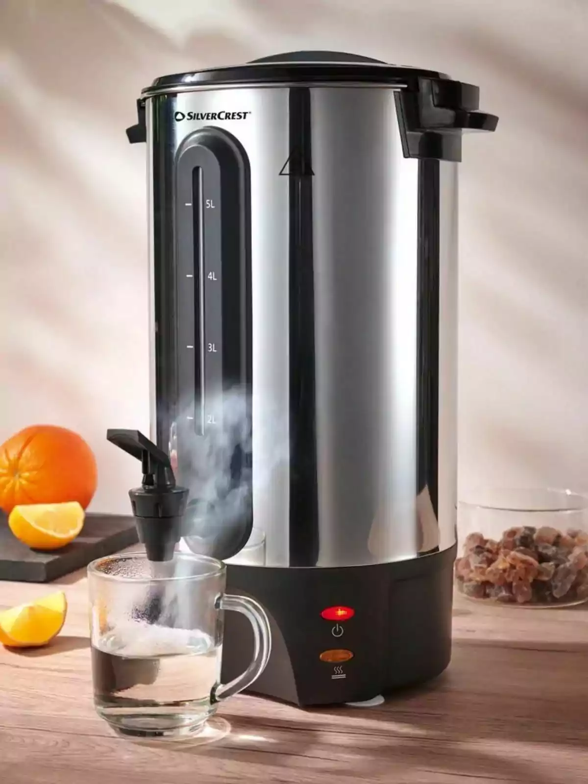 Dispensador d’aigua calenta d’acer inoxidable servint aigua en una tassa de vidre sobre una taula de fusta amb grills de taronja i un recipient amb sucre al fons.