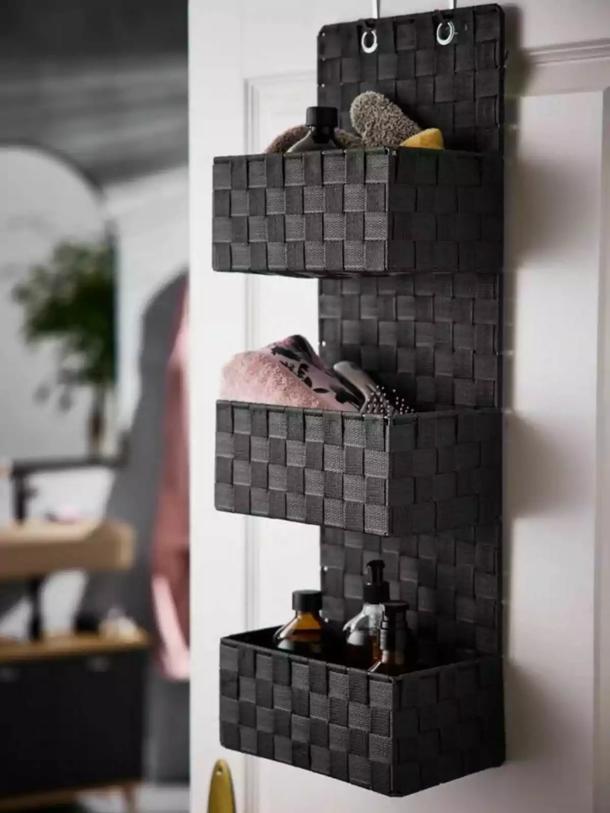 Organitzador de tela negra penjat a una porta amb tres compartiments que contenen ampolles, tovalloles i raspalls.