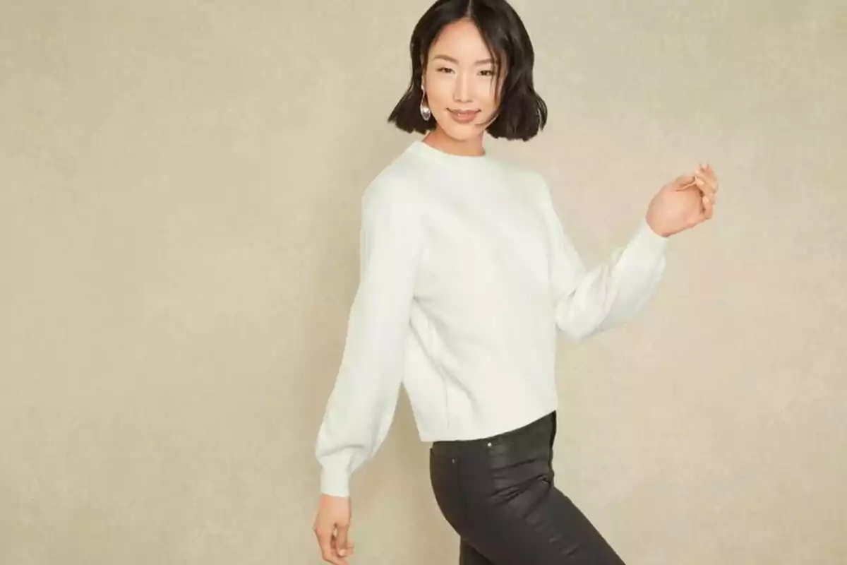 Dona jove amb cabell curt i fosc portant un jersei blanc i pantalons negres posant davant d’un fons beix