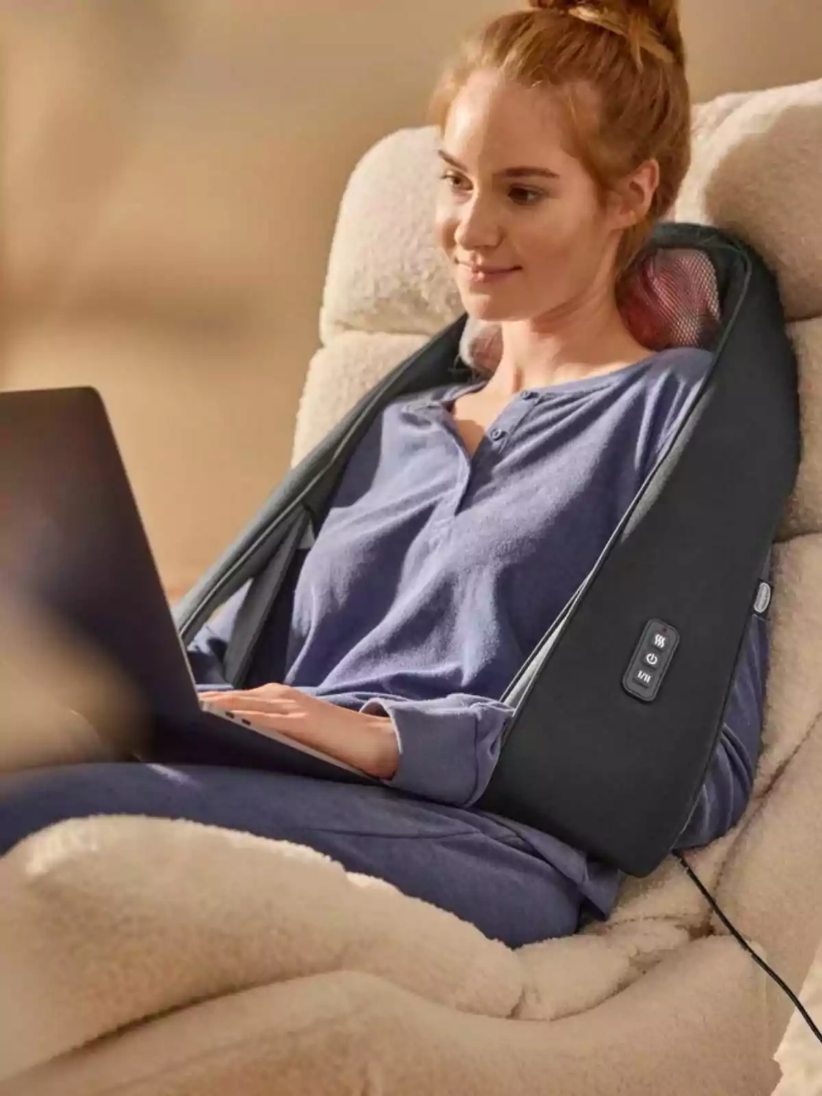 Woman sitting in an armchair using a laptop while enjoying an electric neck and shoulder massager