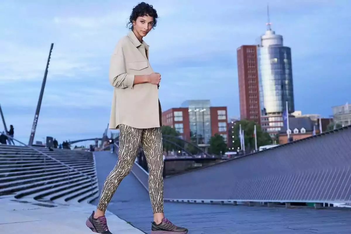 Young woman in a beige jacket and patterned leggings walking through a modern city at sunset