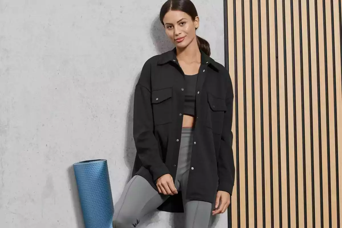 Woman in sportswear and a black jacket leaning against the wall next to a blue yoga mat